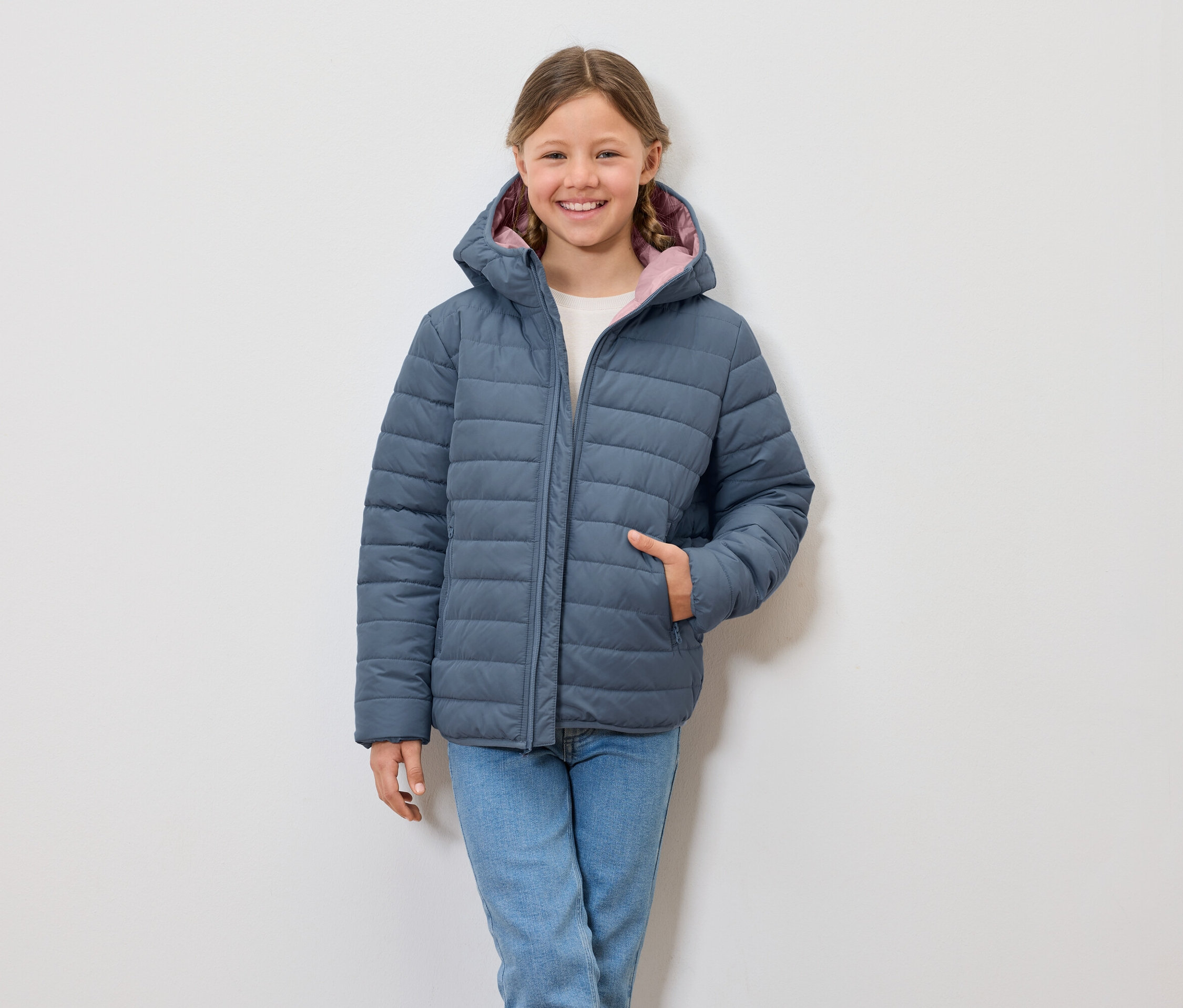 Uśmiechnięta dziewczyna ma na sobie niebieską kurtkę puchową z kapturem i dżinsy.