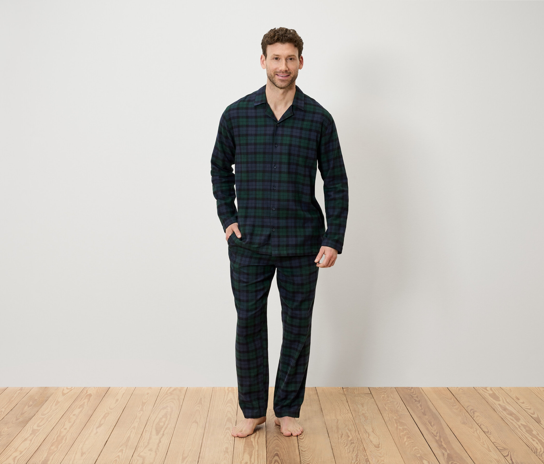 Mężczyzna w piżamie w kratę stoi na drewnianej podłodze przed białą ścianą.