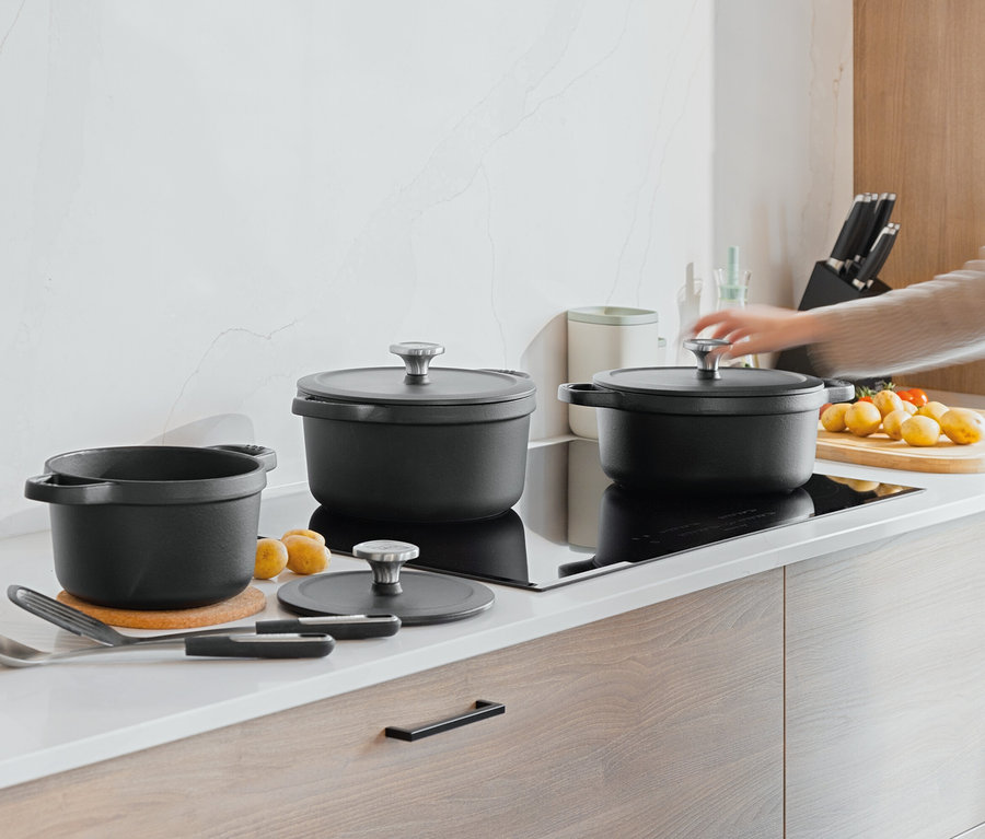 Na blacie kuchennym stoją trzy czarne garnki, obok nich leżą ziemniaki i pomidory. Ręka sięga po pokrywkę jednego z garnków.