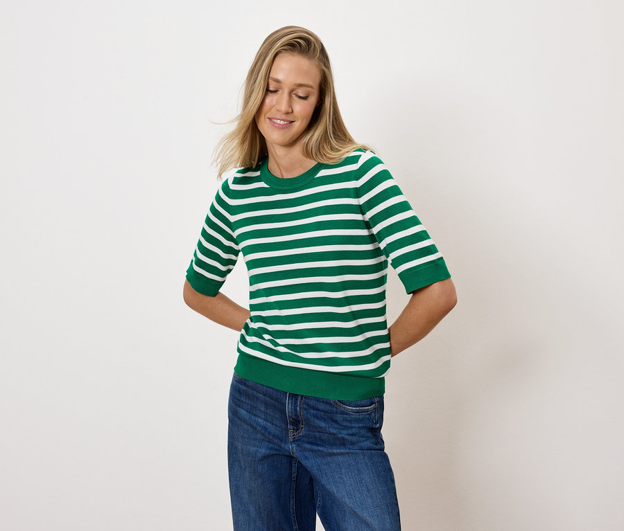 Blondwłosa kobieta pozuje w zielono-białej bluzce w paski i niebieskich dżinsach.