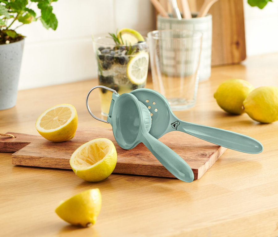 Niebieska wyciskarka do cytryn leży na drewnianej desce obok przekrojonych cytryn. W tle szklanka z cytryną i jagodami.