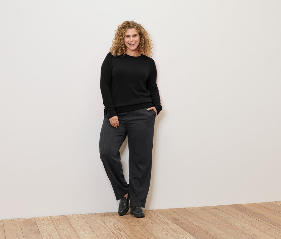 Kobieta o kręconych włosach opiera się o ścianę, ubrana w czarny sweter i wzorzyste spodnie.