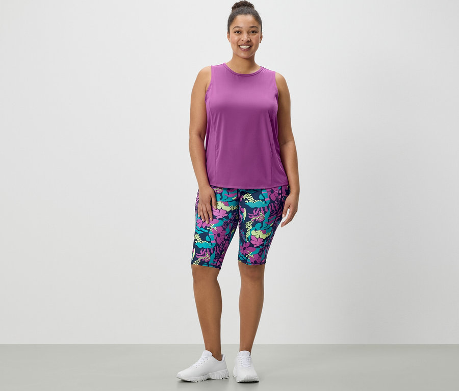 Kobieta stoi i ma na sobie krótkie legginsy z nadrukiem na całej powierzchni oraz turkusowy top sportowy z 2-częściowego zestawu.