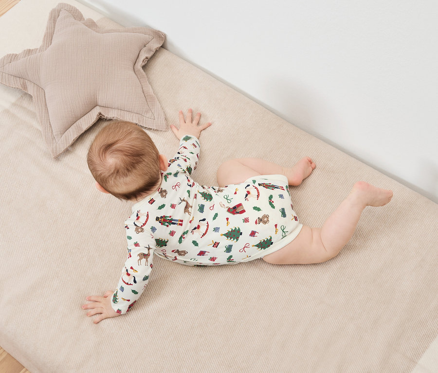 Dziecko leży na brzuchu na kocu z motywem świątecznym obok beżowej poduszki w kształcie gwiazdy.