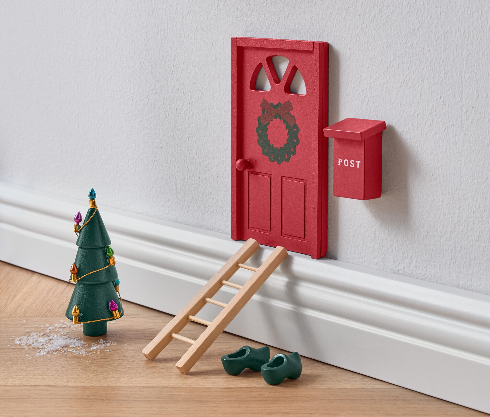 Miniaturowe czerwone drzwi ozdobione wieńcem z drabiną, zielonymi drewniakami i choinką.