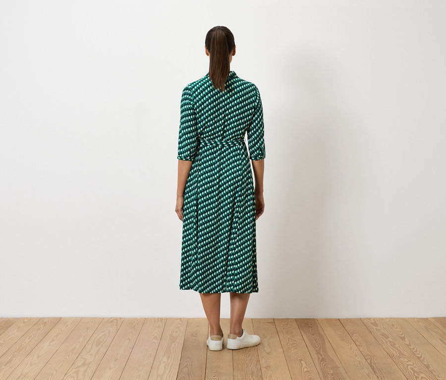 Kobieta stoi tyłem do kamery, ubrana w zieloną sukienkę w białe wzory i białe tenisówki.