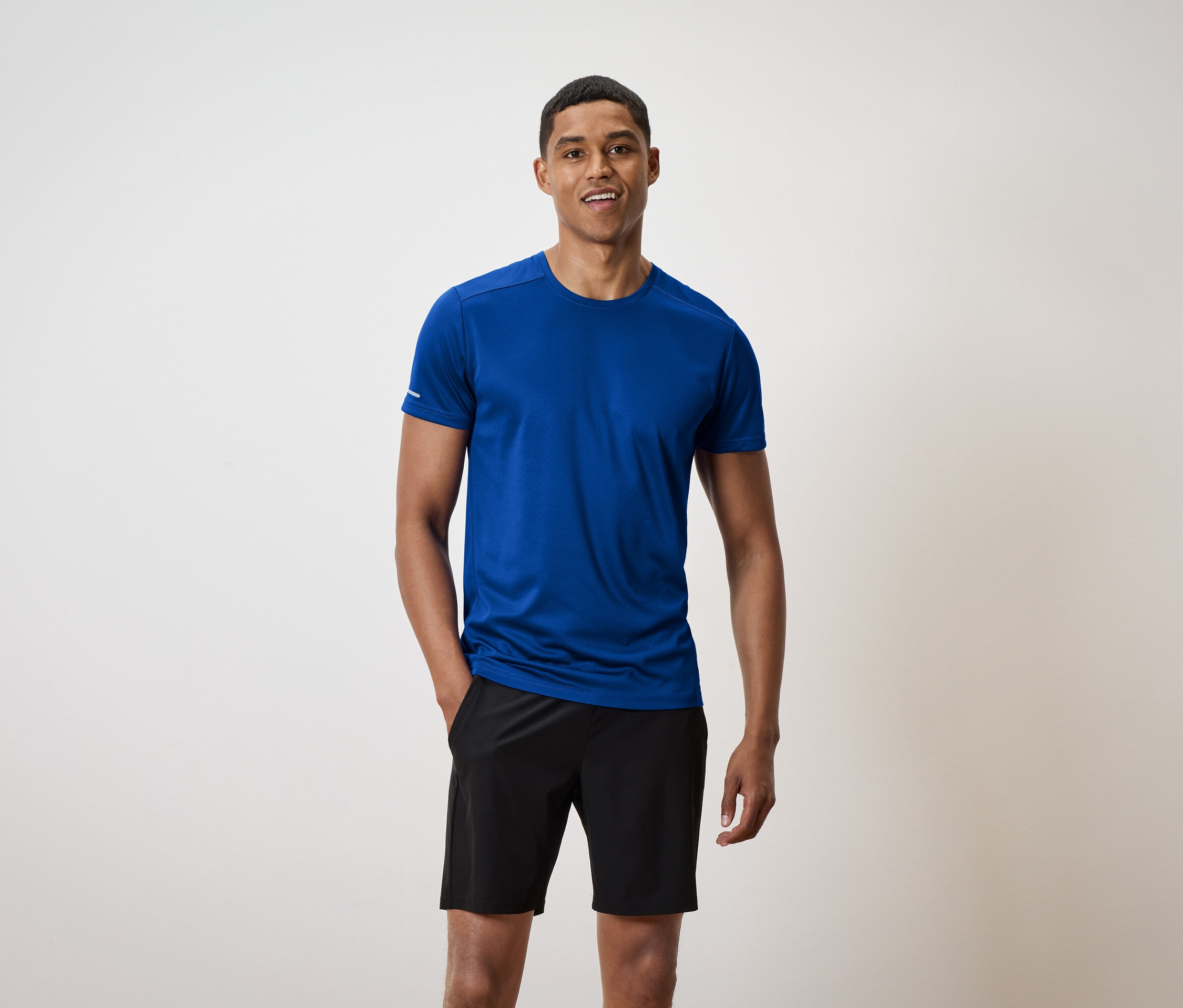 Mężczyzna z krótkimi włosami stoi i uśmiecha się, ma na sobie niebieską koszulkę i czarne spodenki.