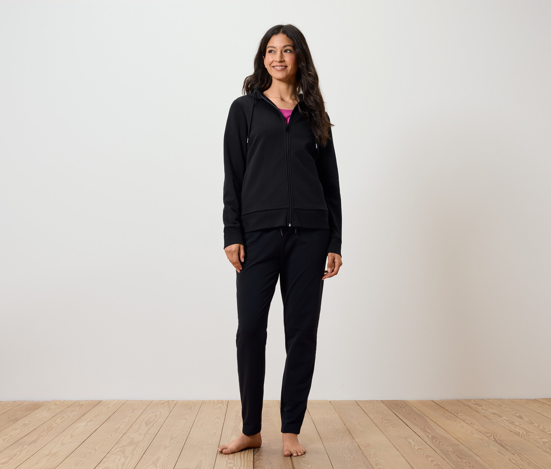 Uśmiechnięta kobieta stoi boso na drewnianej podłodze. Ma na sobie czarne spodnie i czarną bluzę z kapturem zapinaną na zamek.