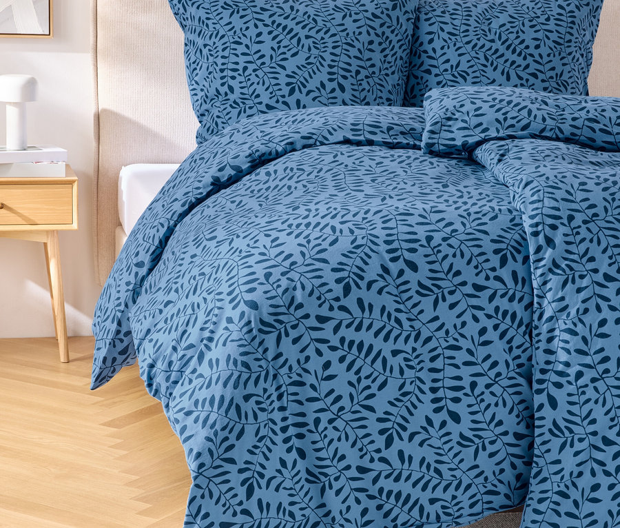 Niebieski zestaw pościeli z wzorem liści na łóżku z zagłówkiem. Obok łóżka stoi stolik nocny z lampką.