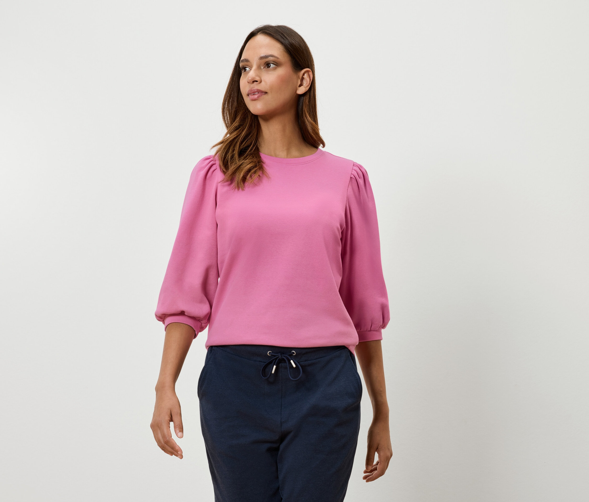 Kobieta ma na sobie różową bluzę z rękawami typu balon o długości trzy czwarte i granatowe spodnie.