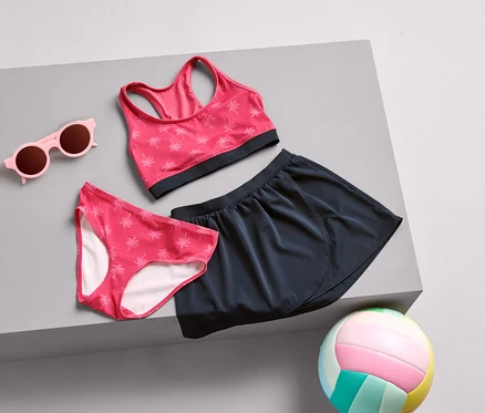 Różowe okulary przeciwsłoneczne, majtki od bikini w palmy, stanik sportowy i granatowa spódnica leżą na szarej powierzchni, obok kolorowa piłka do siatkówki.