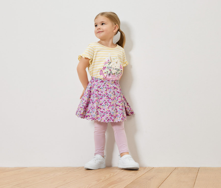 Mała blondwłosa dziewczynka ma na sobie żółto-pasiastą koszulkę z kwiatowym wzorem, kwiecistą spódniczkę, różowe legginsy i białe tenisówki.