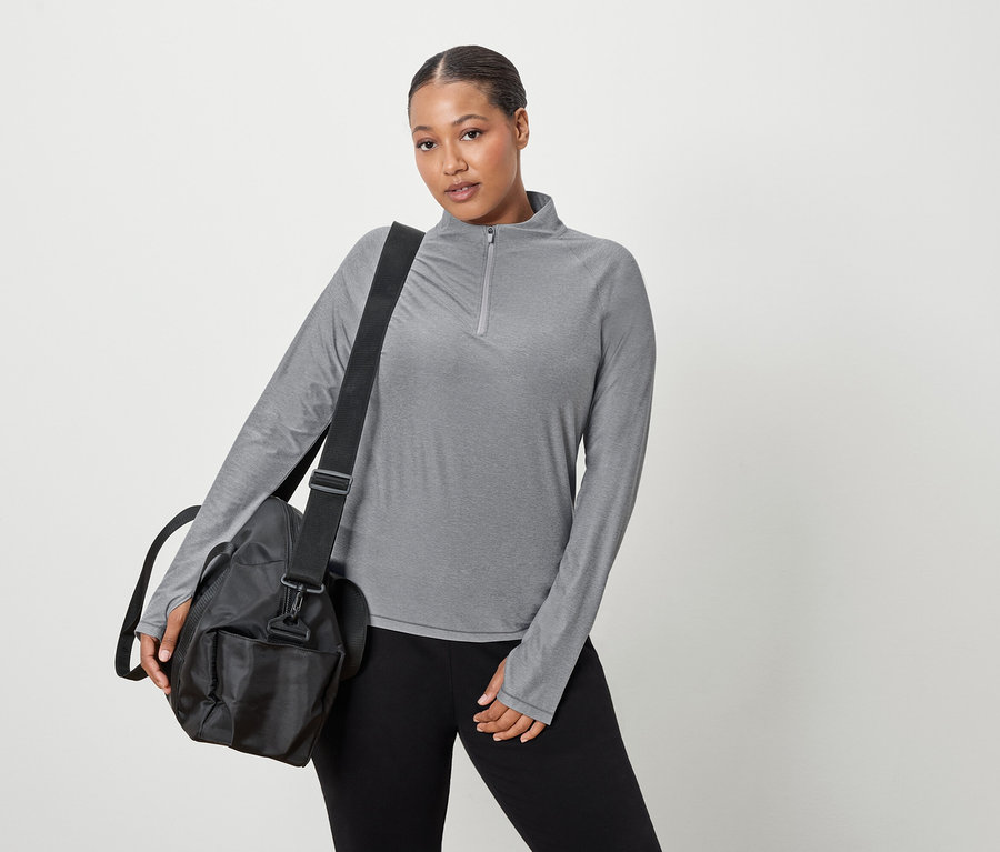 Kobieta ma na sobie szarą bluzkę z długim rękawem i czarną torbę sportową na ramieniu.