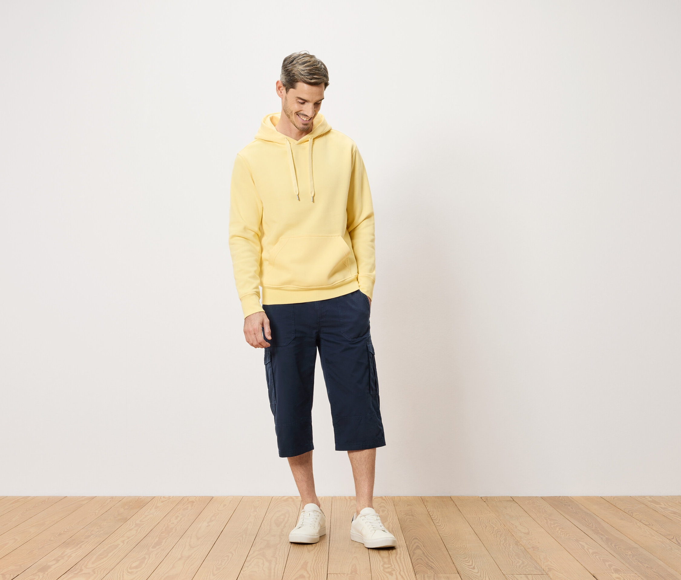Mężczyzna w żółtej bluzie z kapturem i niebieskich szortach cargo stojący na drewnianej podłodze.