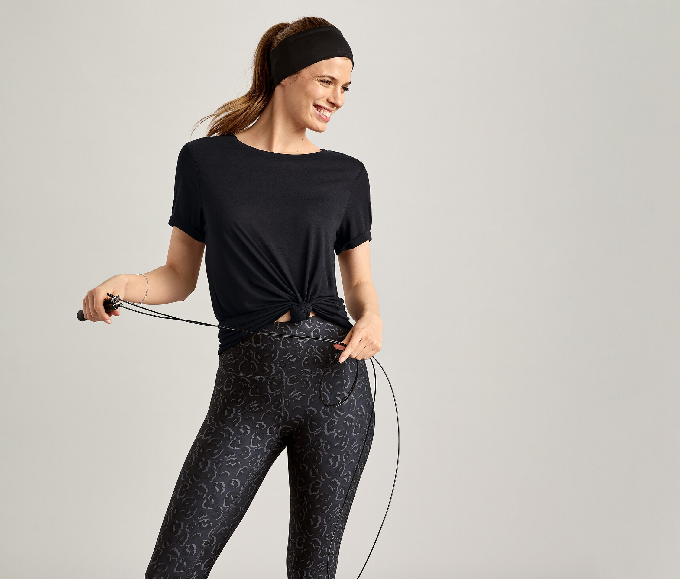Uśmiechnięta kobieta w czarnej koszulce sportowej trzyma skakankę na szarym tle.
