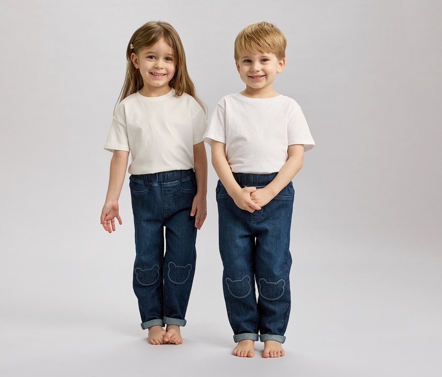 Dwoje dzieci z blond i brązowymi włosami stoi obok siebie boso, ubrane w białe T-shirty i niebieskie dżinsy z motywami niedźwiedzi.