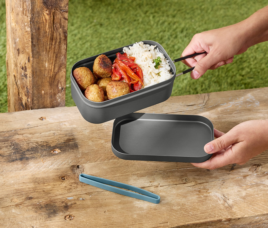 Ręce trzymają szare pudełko na lunch z ryżem, klopsikami i papryką nad szarą pokrywką na drewnianym stole.