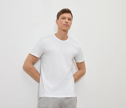 Mężczyzna opiera się o białą ścianę, ma na sobie biały T-shirt i szare spodnie dresowe.