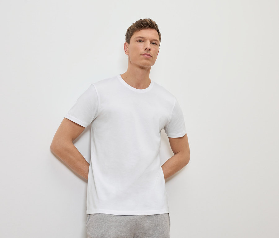 Mężczyzna opiera się o białą ścianę, ma na sobie biały T-shirt i szare spodnie dresowe.