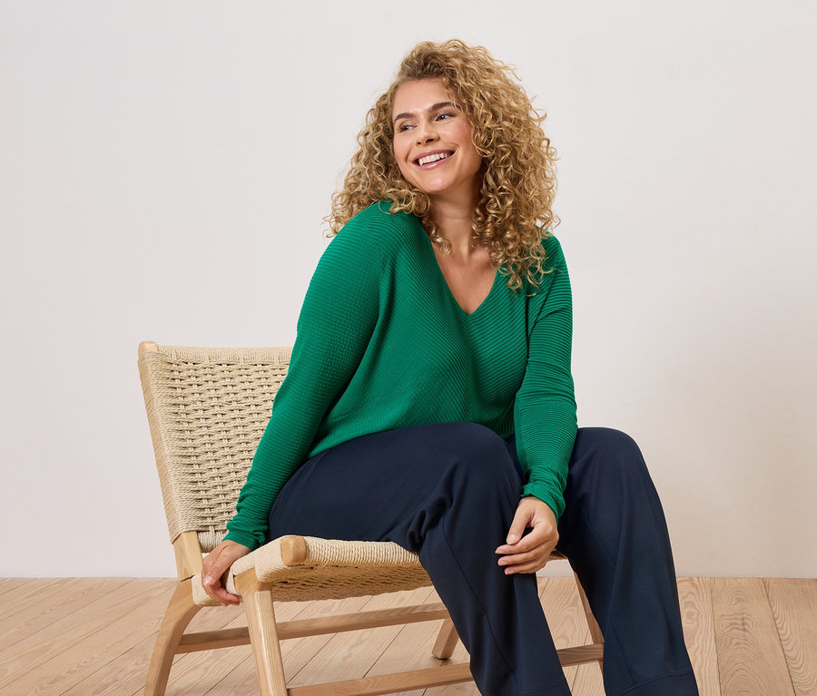 Kobieta o kręconych włosach siedzi na krześle, ubrana w zielony sweter i niebieskie spodnie.