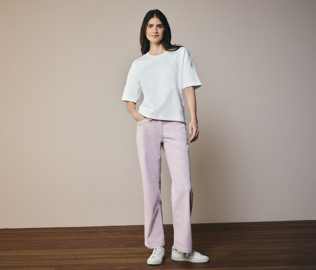 Kobieta w białej koszulce i dżinsach CECIL „Neele”. Stoi na drewnianej podłodze na beżowym tle.