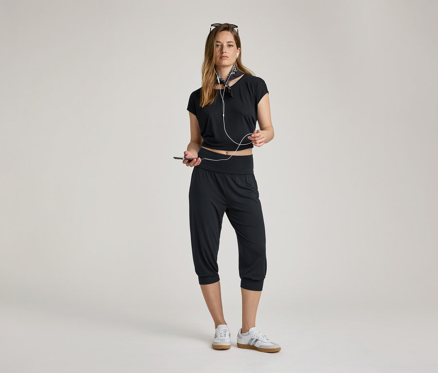 Kobieta ze słuchawkami i telefonem. Nosi spodnie do jogi o długości 3/4, czarny top i tenisówki.