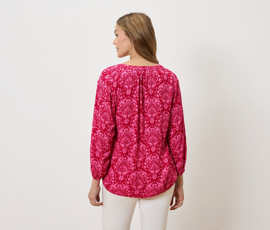 Widok z tyłu kobiety z blond włosami, ubranej w różową bluzkę z długim rękawem we wzory i białe spodnie.