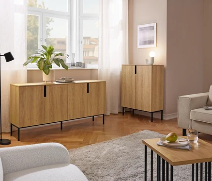 Przytulny salon z długą drewnianą komodą pod oknem i pasującą wysoką szafką. Widoczne są również czarna lampa podłogowa, dwa fotele, kudłaty dywan i stolik kawowy z gruszką i szklanką wody.
