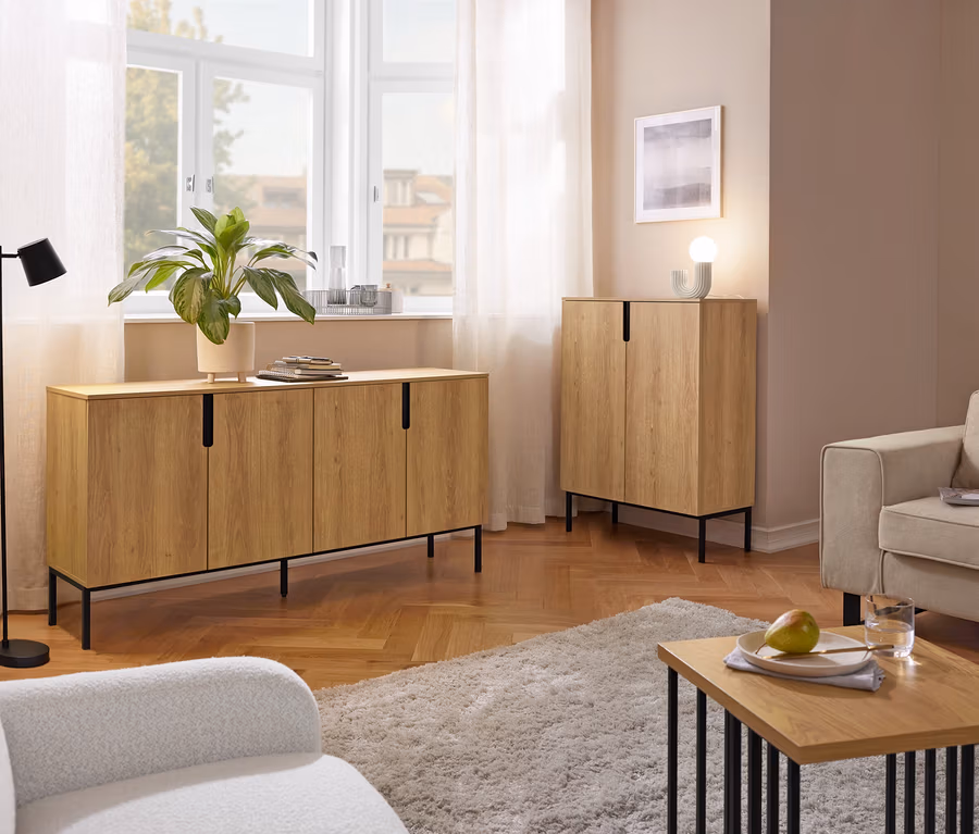 Przytulny salon z długą drewnianą komodą pod oknem i pasującą wysoką szafką. Widoczne są również czarna lampa podłogowa, dwa fotele, kudłaty dywan i stolik kawowy z gruszką i szklanką wody.
