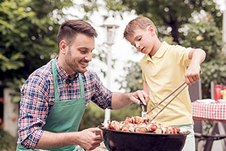 Młody mężczyzna w zielonym fartuchu kuca przy czarnym okrągłym grillu, a jego syn, trzymając szczypce, obraca szaszłyki.