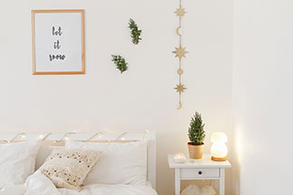 Lampy z jakością Tchibo: utrzymana w tonacji bieli sypialnia z łóżkiem i stoliczkiem nocnym, na którym stoi włączona dekoracyjna lampka w kształcie grzybka.