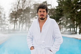 Brodaty mężczyzna w białym szlafroku stoi przed parującym basenem. Ręce mężczyzny skryte są w kieszeniach szlafroka.