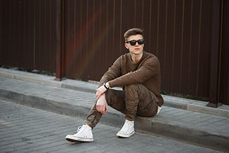 Buty sportowe na co dzień: modnie ubrany młody mężczyzna w sięgających kostki białych sneakersach siedzi na krawężniku.