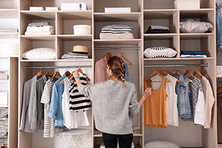 Miejsce do przechowywania dobrej jakości: młoda kobieta stoi przed swoją dużą, otwartą szafą, trzymając w dłoni drewniany wieszak z pasiastym T-shirtem.