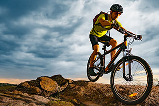 Młody mężczyzna ubrany w strój rowerowy, złożony z trykotu rowerowego z krótkimi rękawami i krótkich spodenek rowerowych, jedzie na rowerze górskim po górzystym terenie.