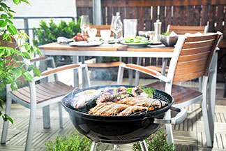 Balkon, na którym stoi grill z mięsem. W tle widać rustykalny, nakryty stół ogrodowy z drewnianymi krzesłami.