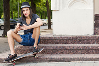 Stylowo ubrany skater w ciemnym kapeluszu i dżinsowych spodenkach siedzi na marmurowych schodach i obsługuje swój smartfon.