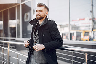 Kurtka męska do biura: młody mężczyzna w czarnym krótkim płaszczu, pod którym ma szarą marynarkę, opiera się o balustradę, trzymając w ręku swoje okulary przeciwsłoneczne.