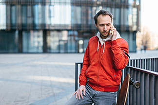 Kurtki sportowe na co dzień: młody mężczyzna w czerwonej kurtce rozmawia przez smartfona, obok niego stoi deskorolka longboard.