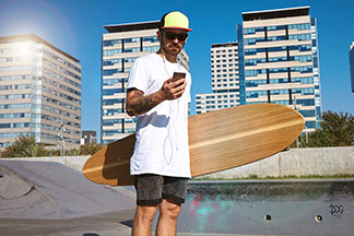 Skater w okularach przeciwsłonecznych, czapce z daszkiem i koszulce oversize, z deskorolką longboard w ręku, stoi w skateparku i patrzy na telefon komórkowy.